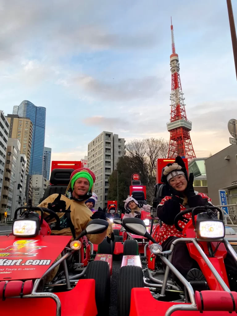 Group of people riding red go-karts on a city street with Tokyo Tower in the background; everyone smiles and wears colorful costumes.