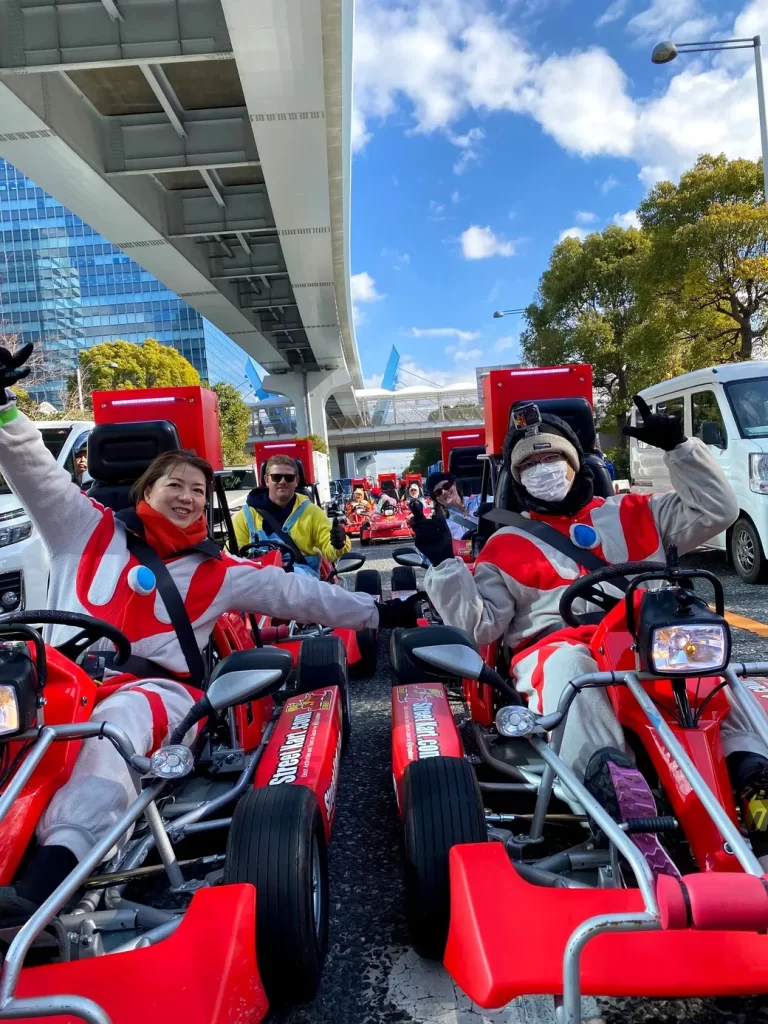 IMG_2083 - StreetKart Group of people in red go-karts on a city street, waving and smiling at the camera under an elevated roadway.